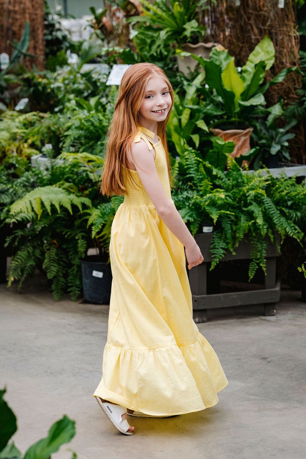 Young person in flowing yellow sundress poses in front of lush green plants and ferns in a garden setting.