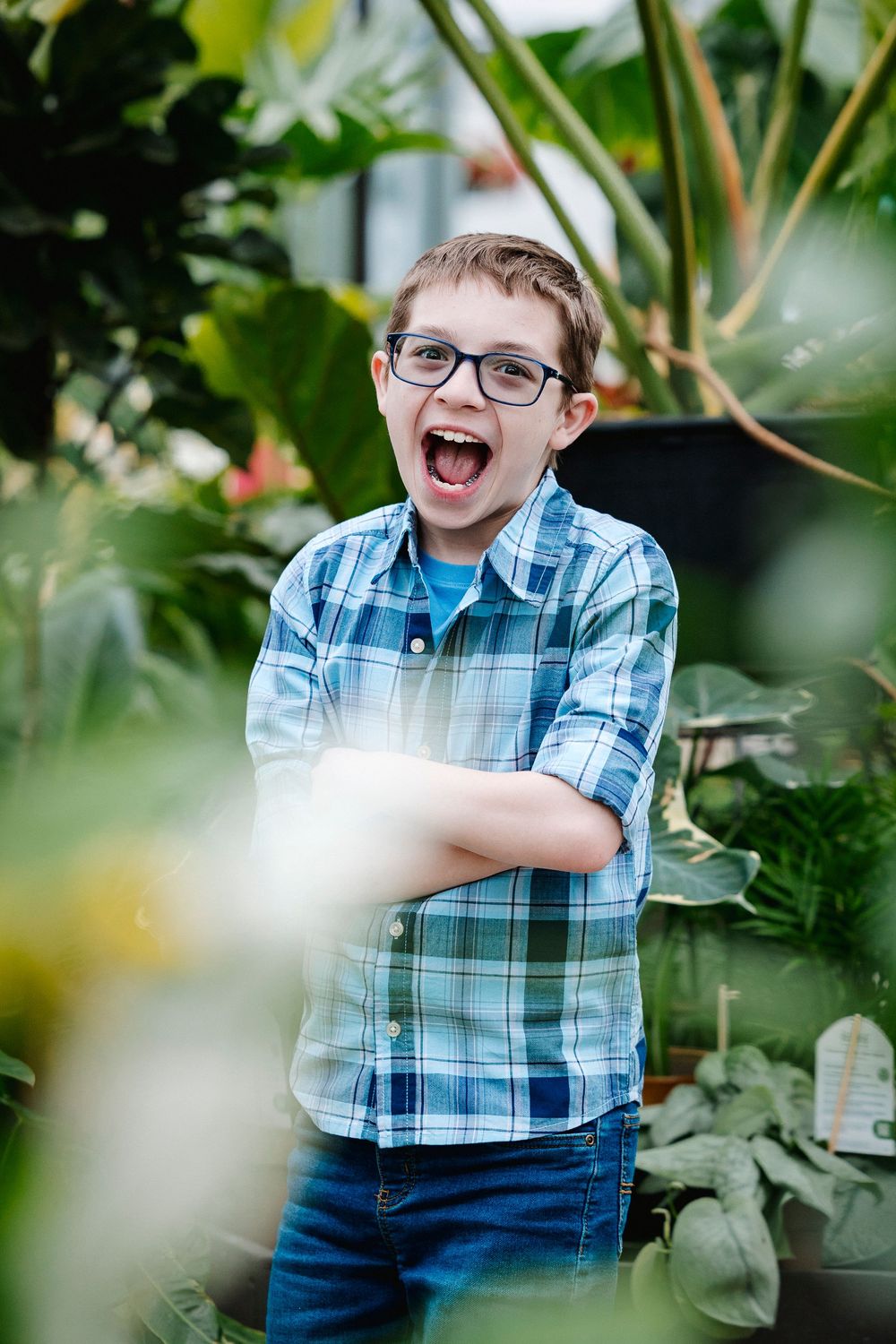 Child wearing blue plaid shirt and glasses stands excitedly among leafy greenery in a garden setting.