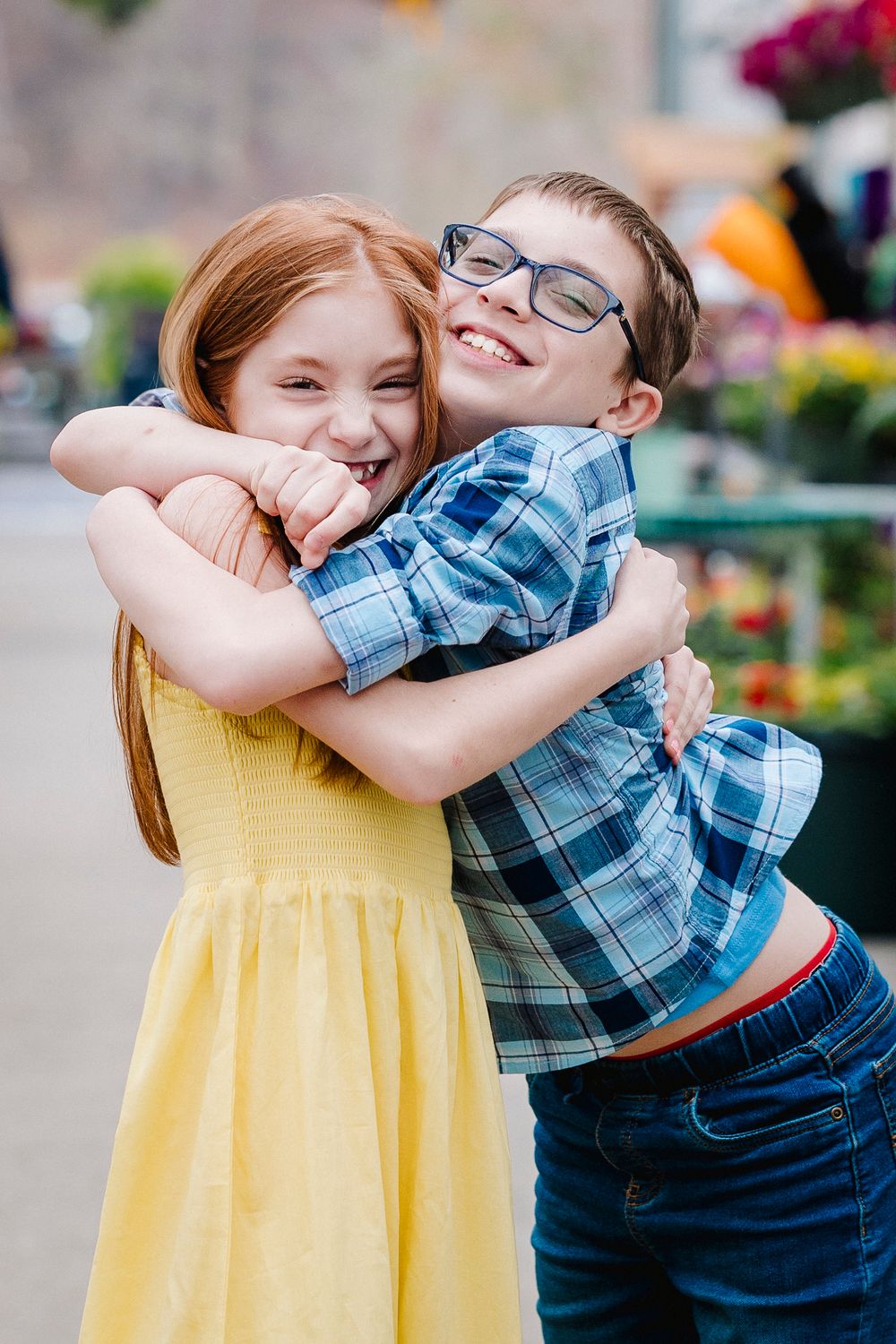 Two youths share a joyful embrace in front of a colorful floral display, one wearing yellow and one in plaid.