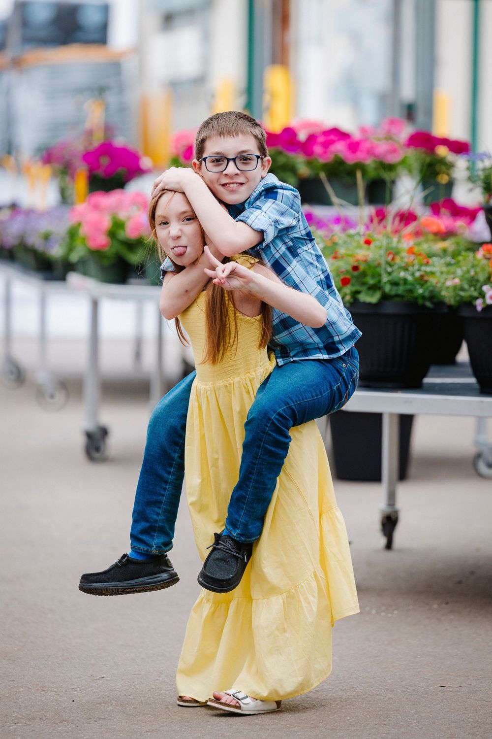 Siblings share a playful moment by colorful flower planters as one gets a piggyback ride in a yellow dress and plaid shirt.