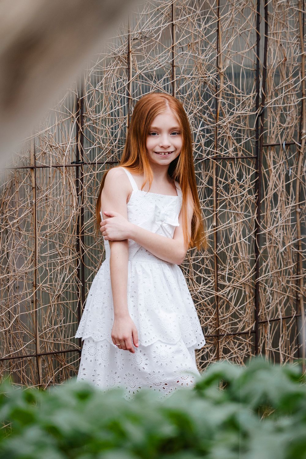 A sweet child in a white lace dress stands outdoors against a backdrop of bare vine-covered branches.