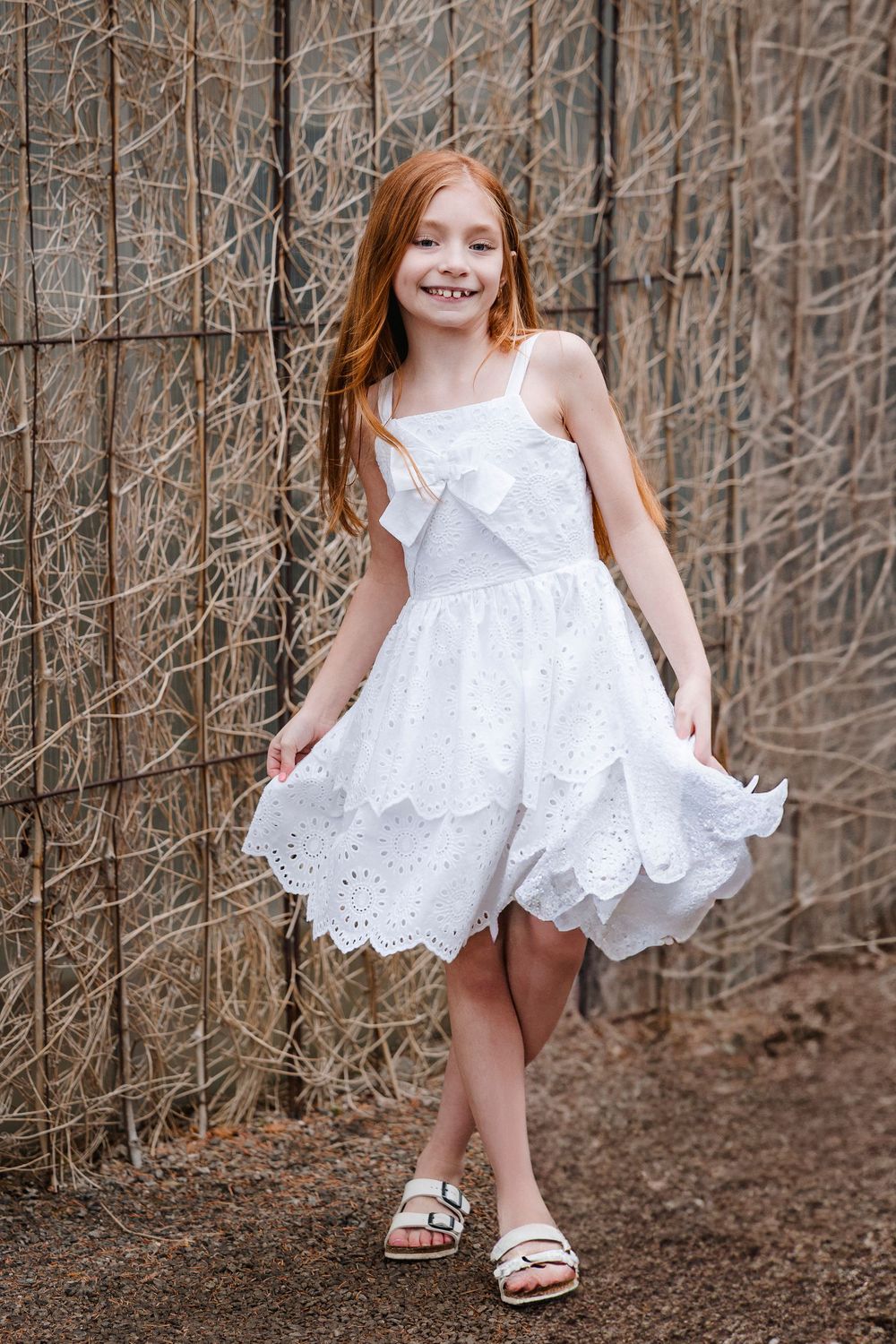 A young girl in white eyelet lace dress twirls playfully against a rustic wall backdrop.