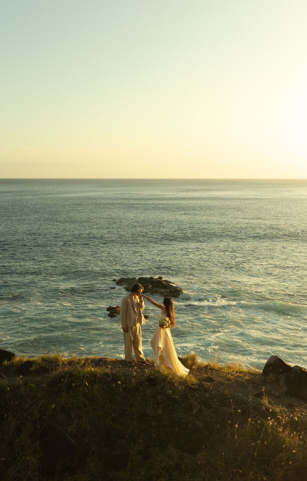 A couple stands at the edge of an ocean cliff at sunset, silhouetted against a dreamy golden sky.