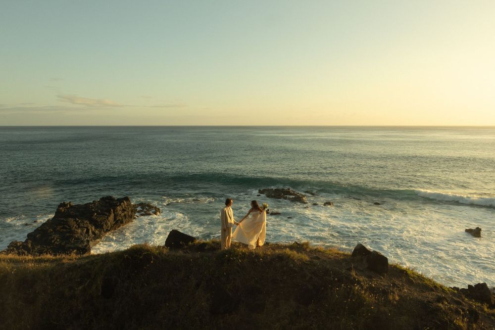 Two silhouettes stand on a rocky cliff overlooking a dramatic ocean coastline at sunset with waves crashing below.