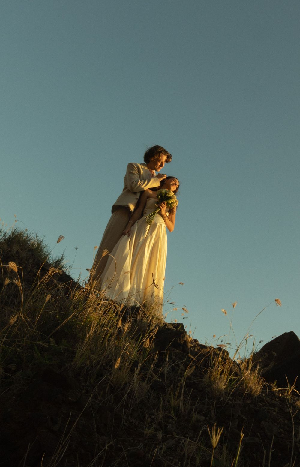 A silhouetted couple in romantic poses stands on a hillside at golden hour against a clear blue sky.