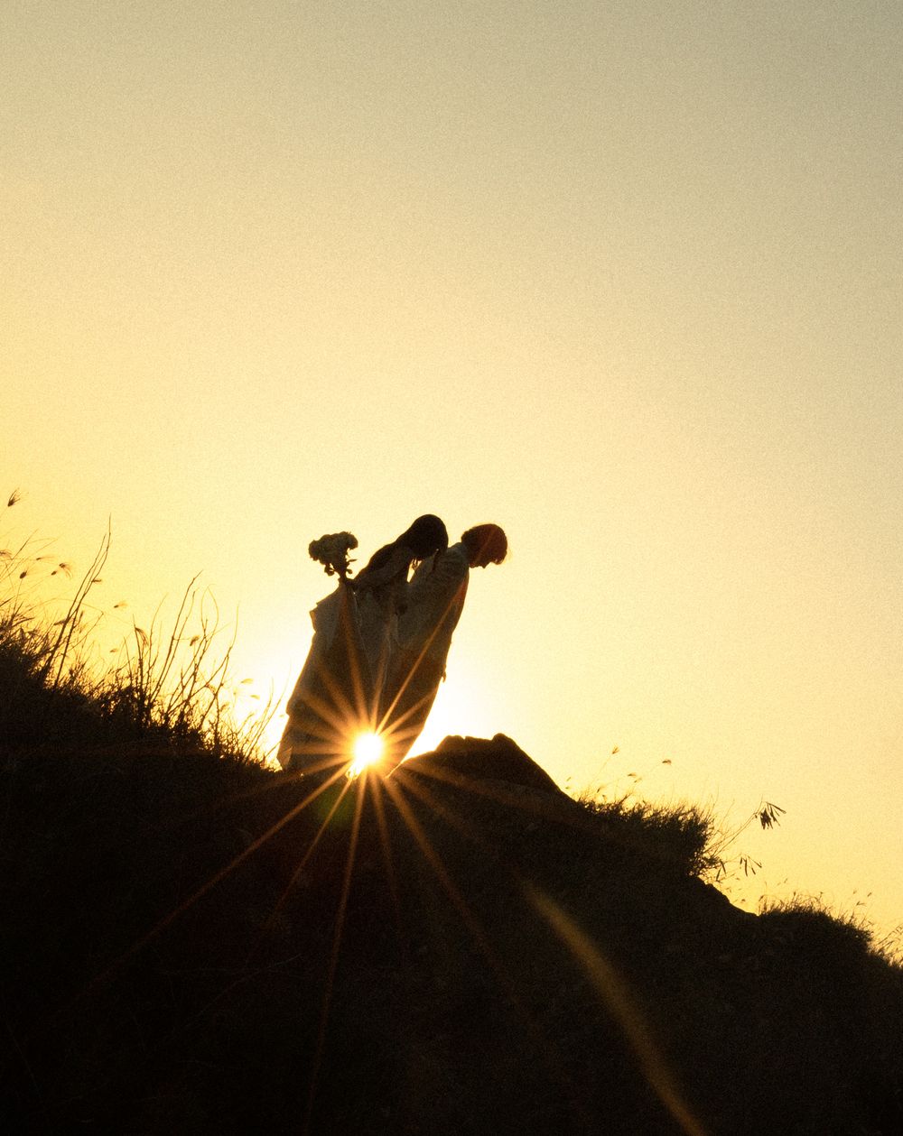 Silhouette of a figure stands on a hillside during golden hour sunset with a dramatic sunflare at their feet.
