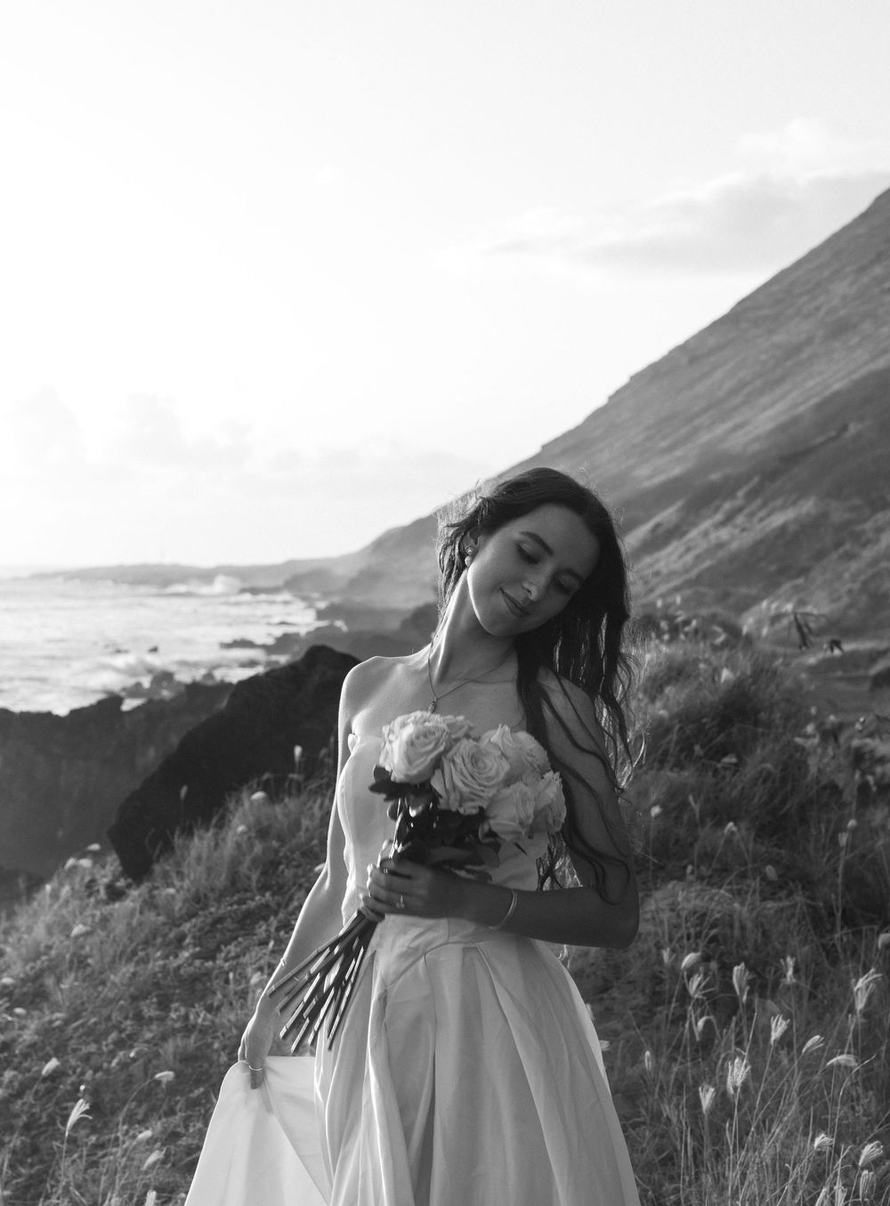 A person in a white dress holds a bouquet while posing against a scenic coastal cliff backdrop in black and white.