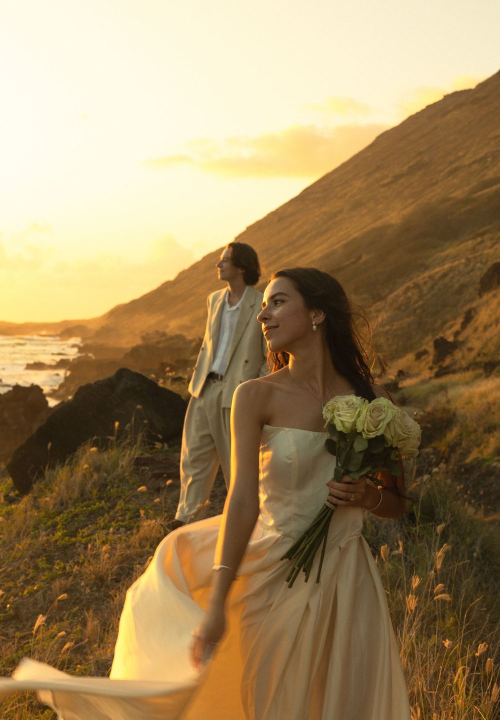 A wedding couple stands on a coastal hillside at golden hour with the ocean and mountains in the background.