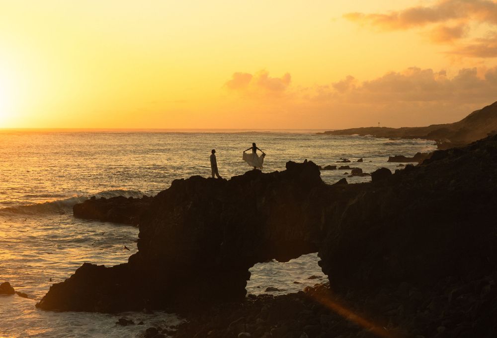 Silhouetted figures enjoy sunset view from rocky cliff overlooking Pacific Ocean waves at Hawaiian coastline.