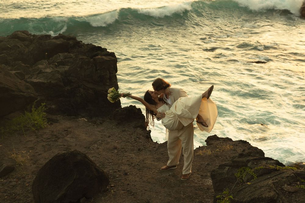 A couple in white attire share a romantic moment on rocky cliffs overlooking dramatic ocean waves at sunset.