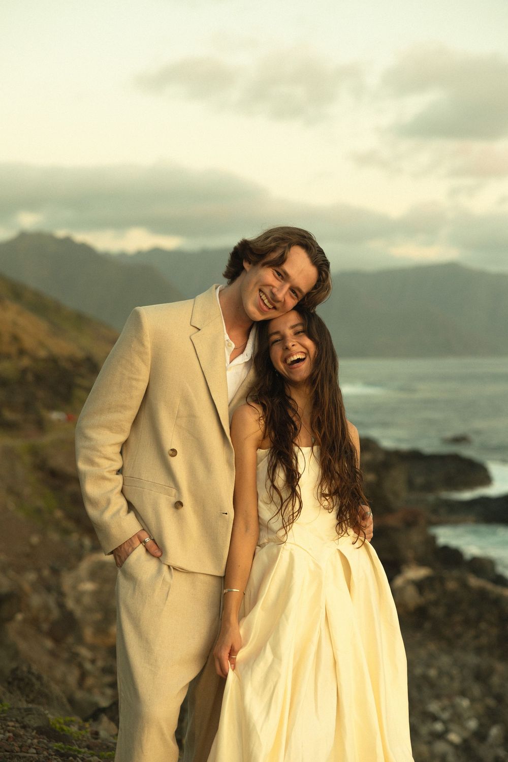 A joyful couple in cream-colored formal attire share a laugh against a dramatic coastal mountain backdrop at sunset.