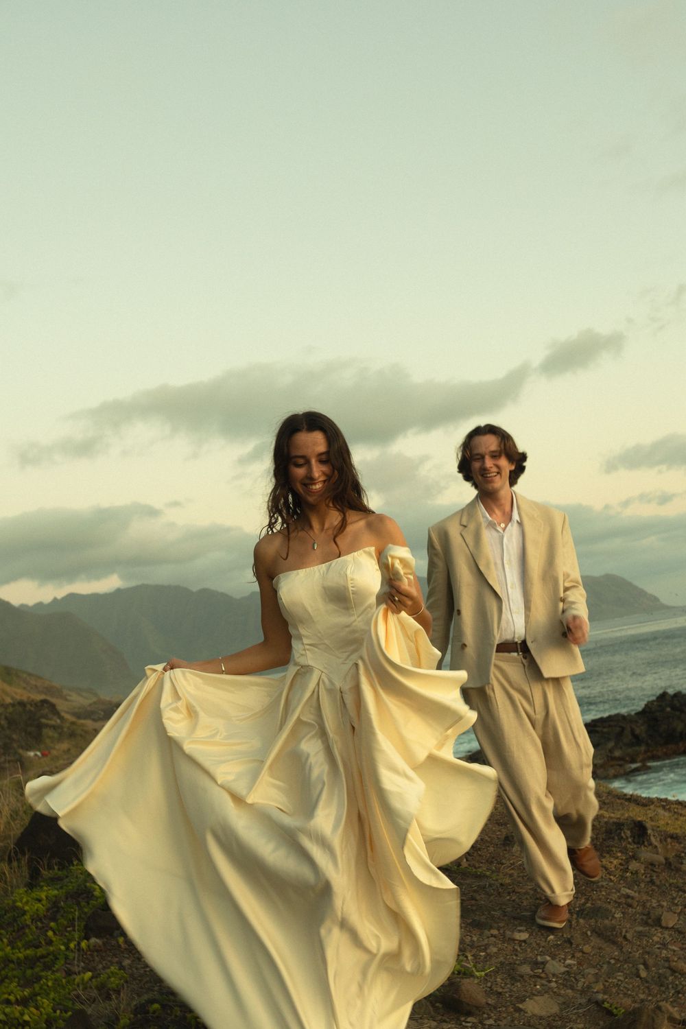 A romantic wedding couple poses on a mountain cliff at sunset, with the bride's ivory dress flowing in the wind.
