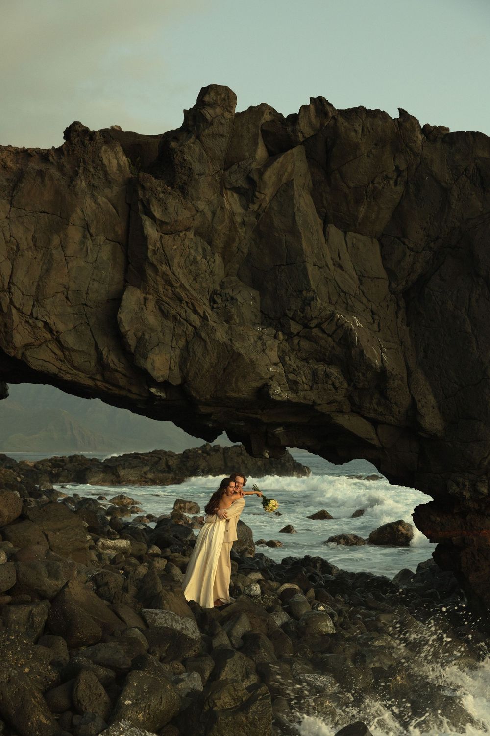 A dreamy sunset photo of a figure in a flowing dress standing beneath a natural rock arch along a rugged coastline.