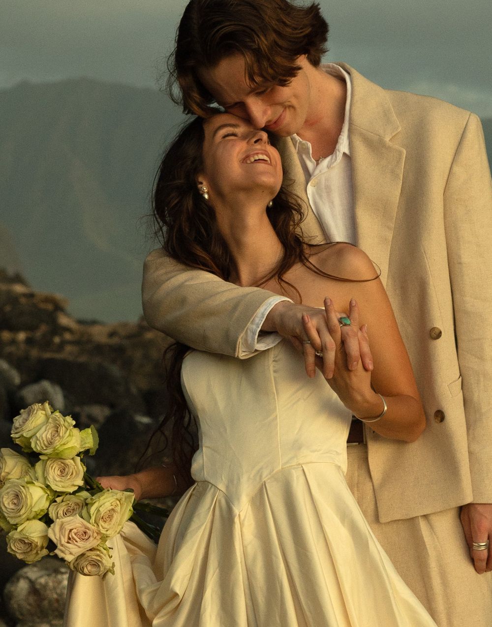 A romantic couple in coordinating cream-colored formal wear embrace on a hillside with yellow roses at sunset.