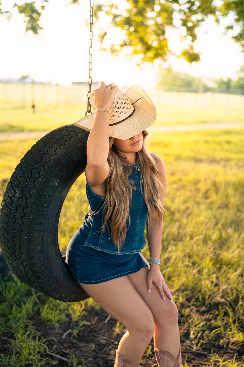 A woman in denim sits on a tire swing in a golden sunlit field wearing a cowboy hat.