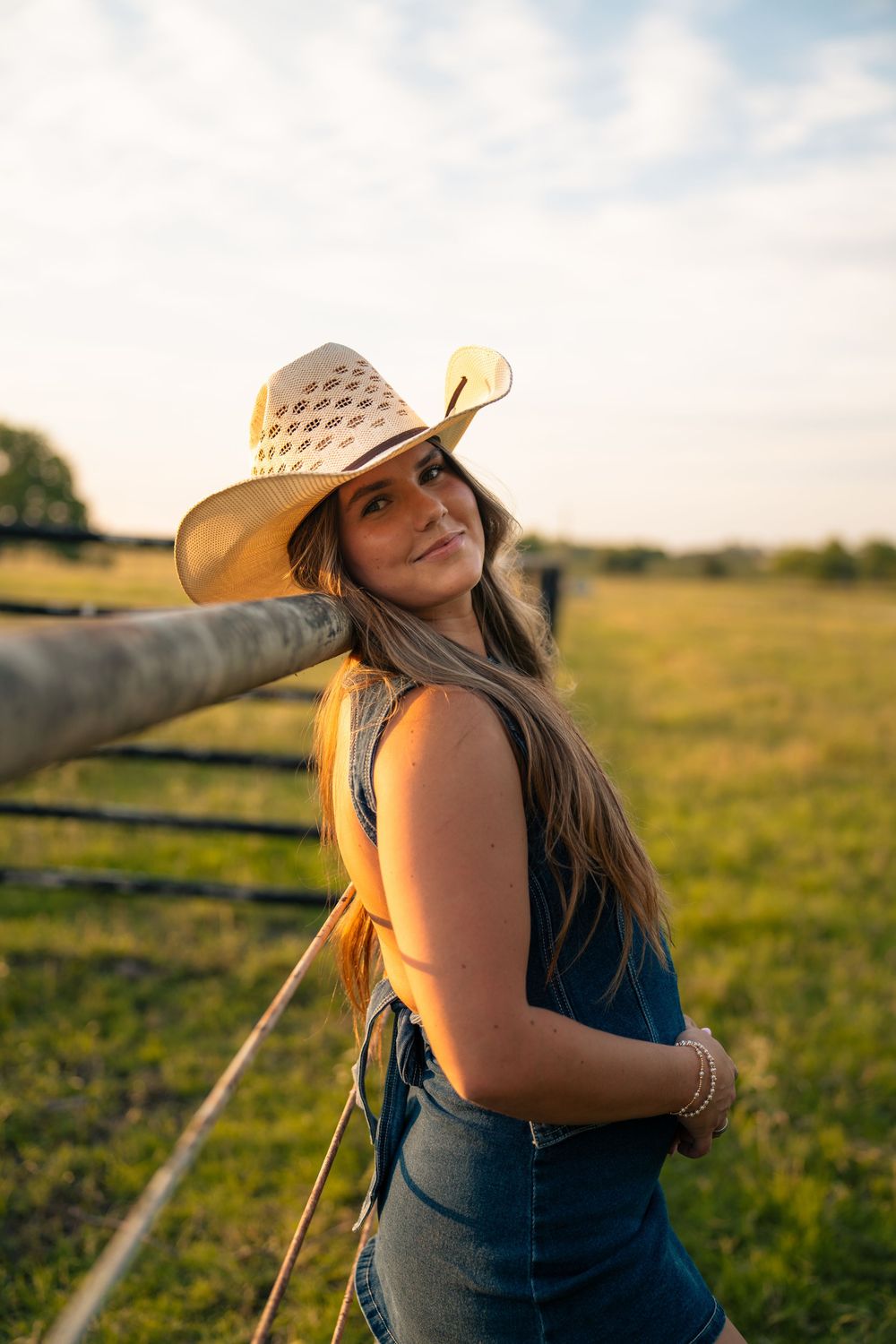 A woman in a denim top and cowboy hat poses by a fence in a sunny field at golden hour.