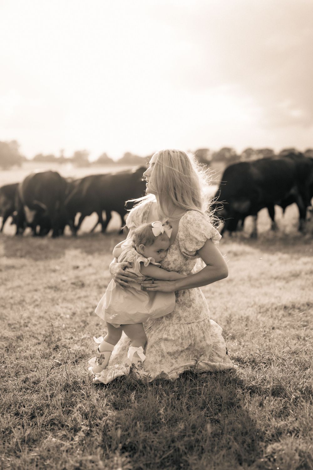 A woman in a flowing vintage dress stands in a field with grazing buffalo during sunset, captured in sepia tones.