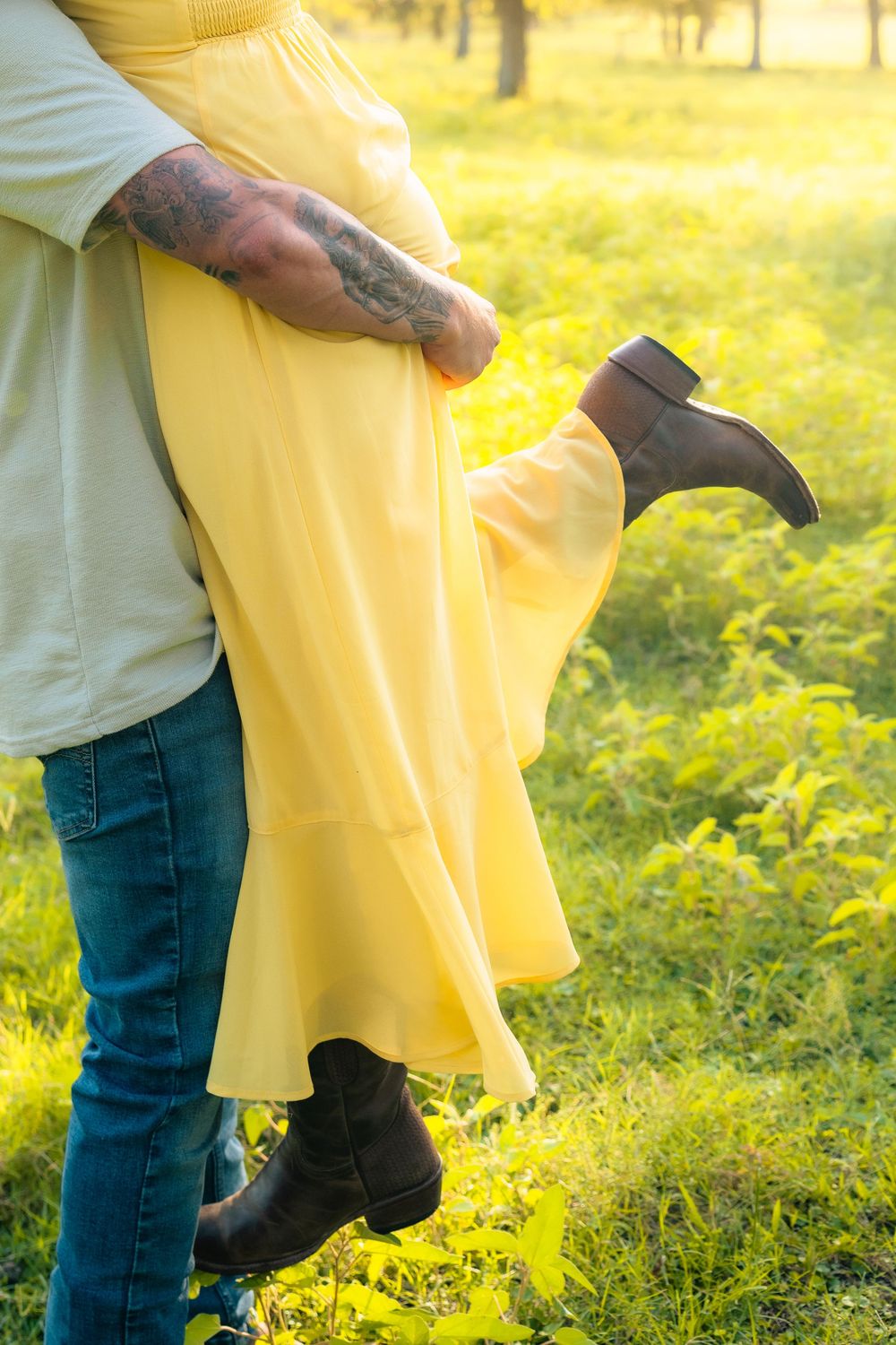 Couple in yellow and beige outfits embracing outdoors in a sunlit grassy field during golden hour.