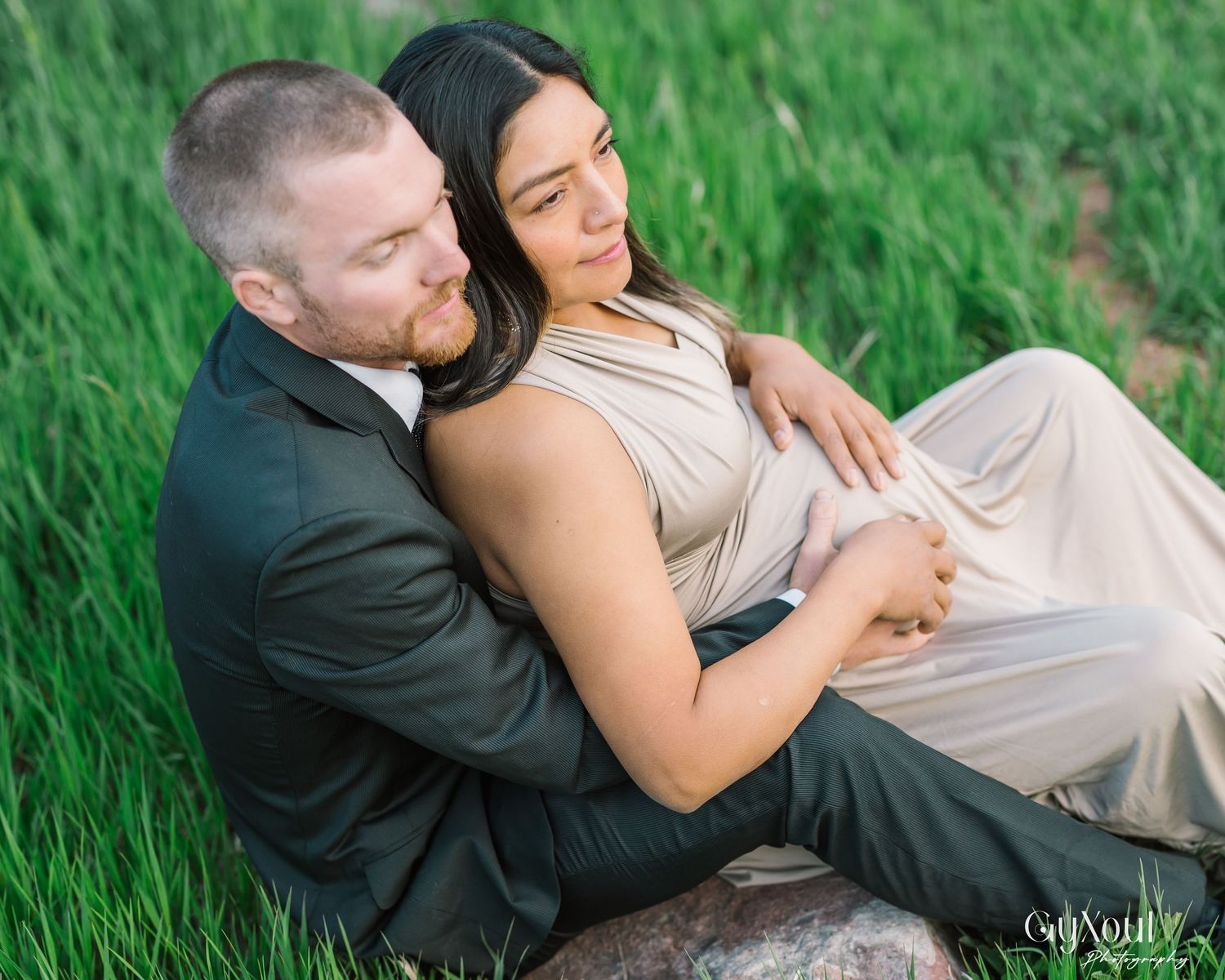 A couple in formal attire embraces while sitting in lush green grass during a romantic outdoor photo session.