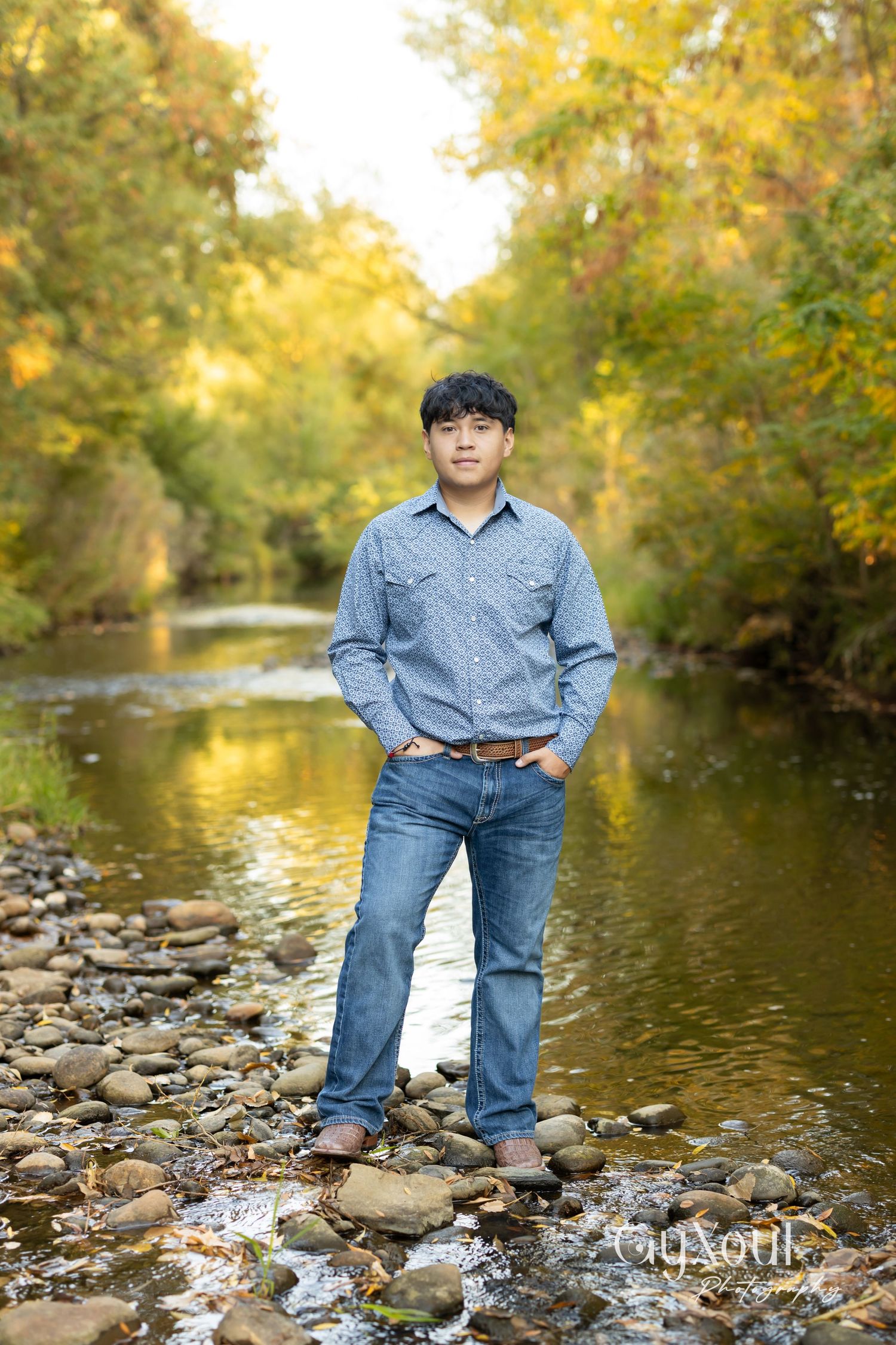 Person stands on rocky riverbank surrounded by autumn trees with golden foliage reflecting in calm water.