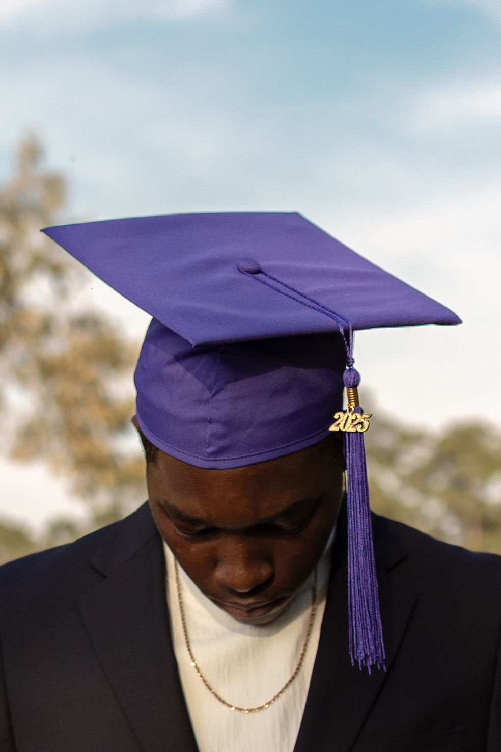 A graduate wears a purple mortarboard cap with gold tassel and black suit against a soft blue sky backdrop.
