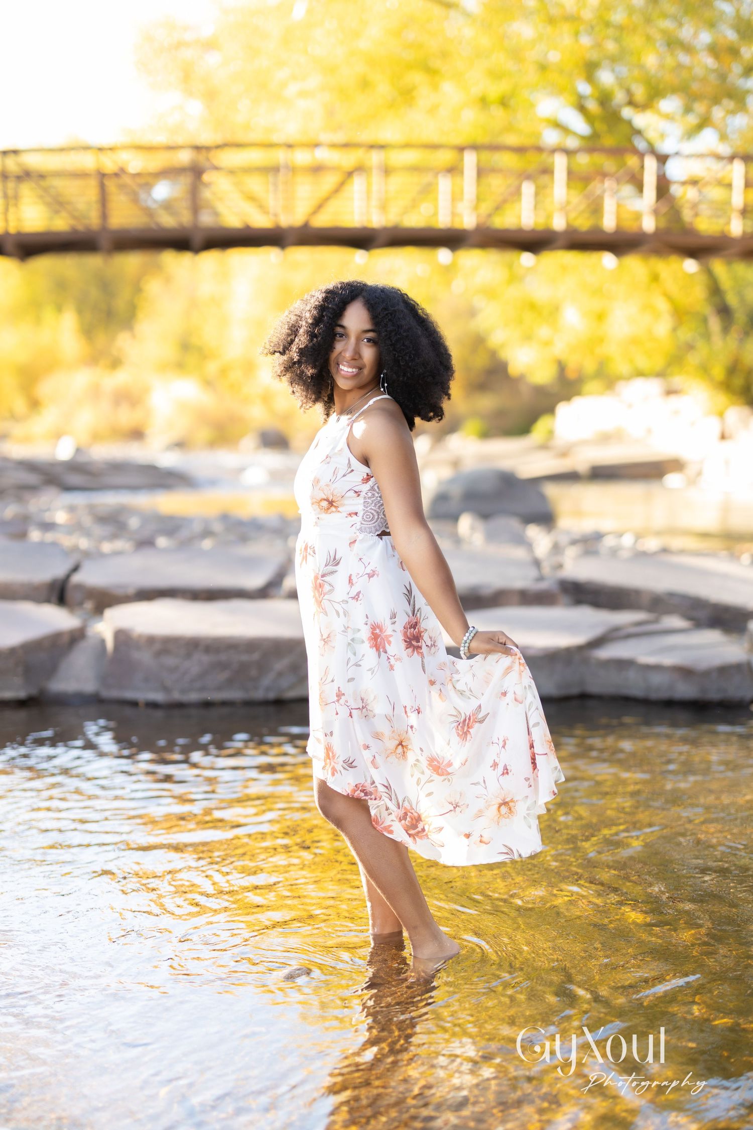 Woman in floral white dress standing in shallow river water with golden autumn foliage and bridge in background.