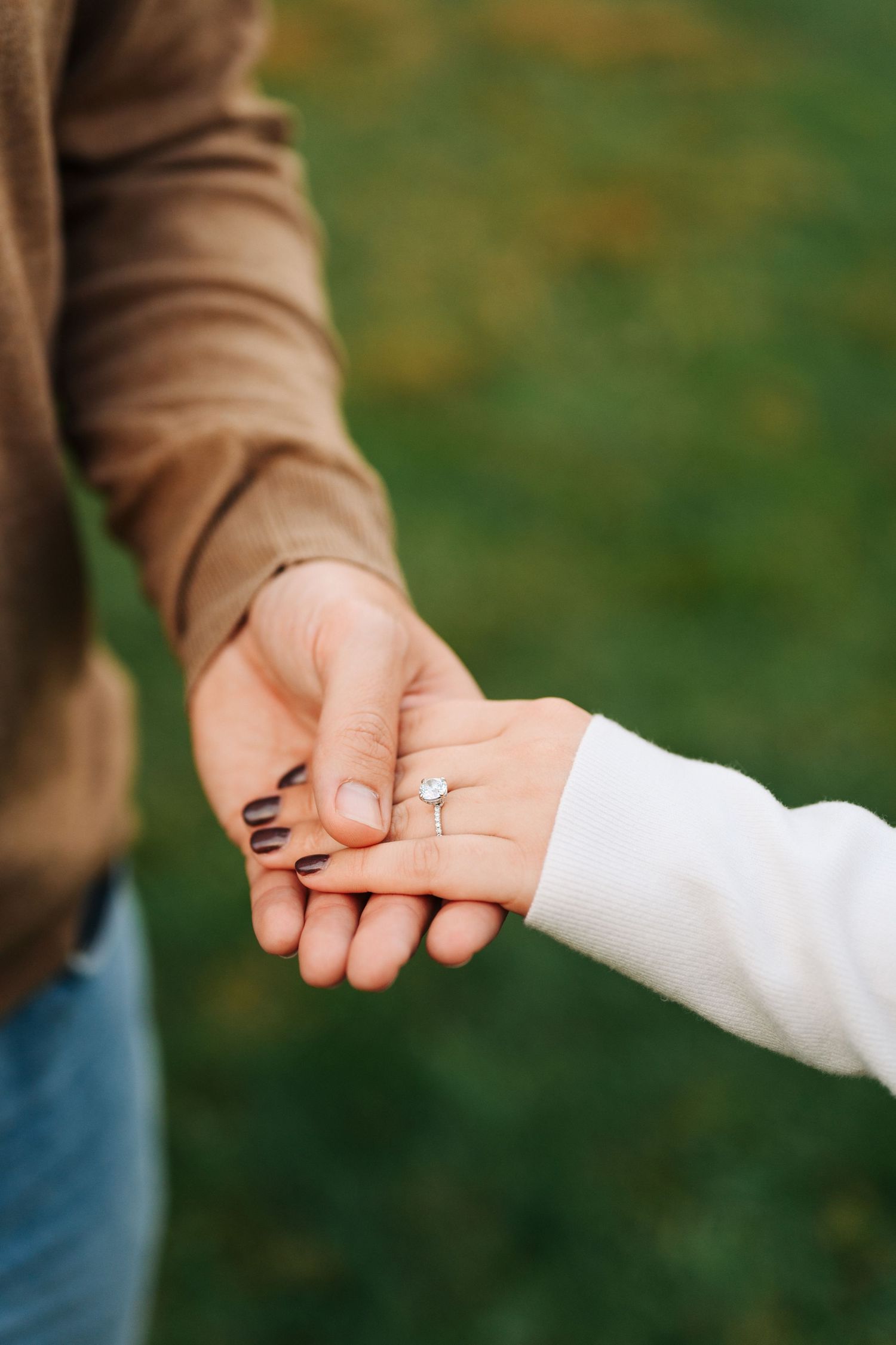 Two hands hold gently together revealing a sparkling diamond engagement ring against a soft blurred green background.