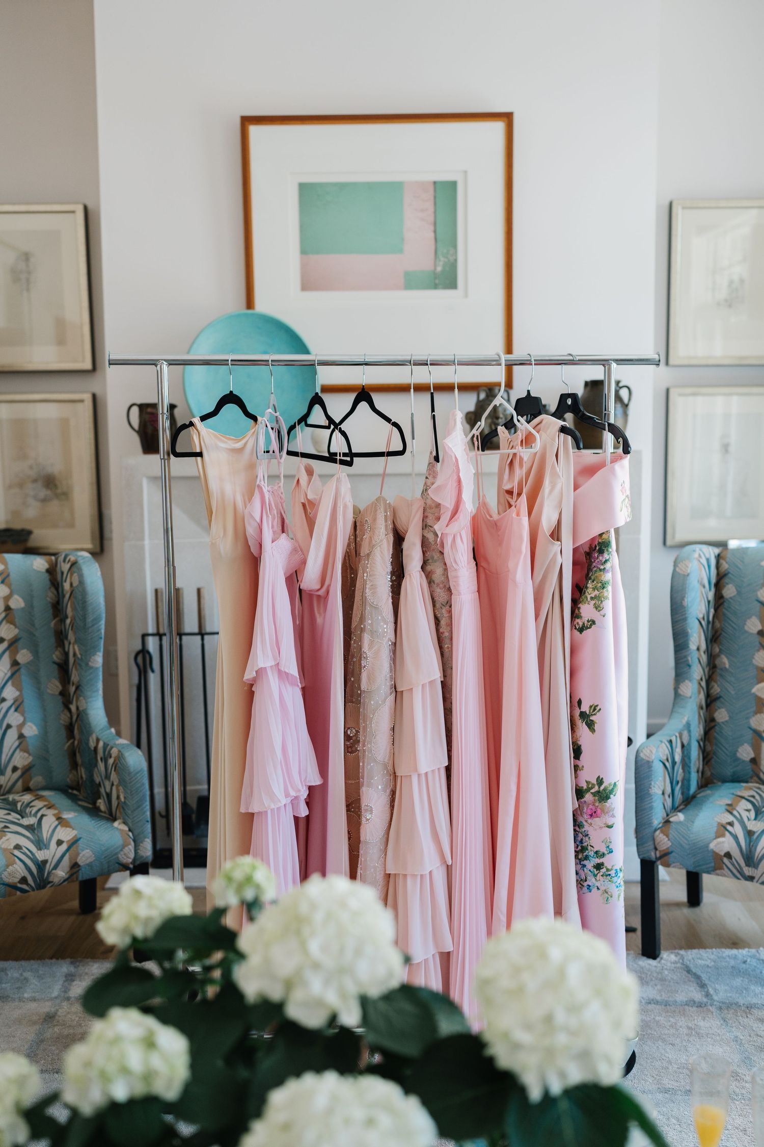 Pink bridesmaid dresses hanging on a rack with turquoise accents and white hydrangeas in an elegant dressing room.