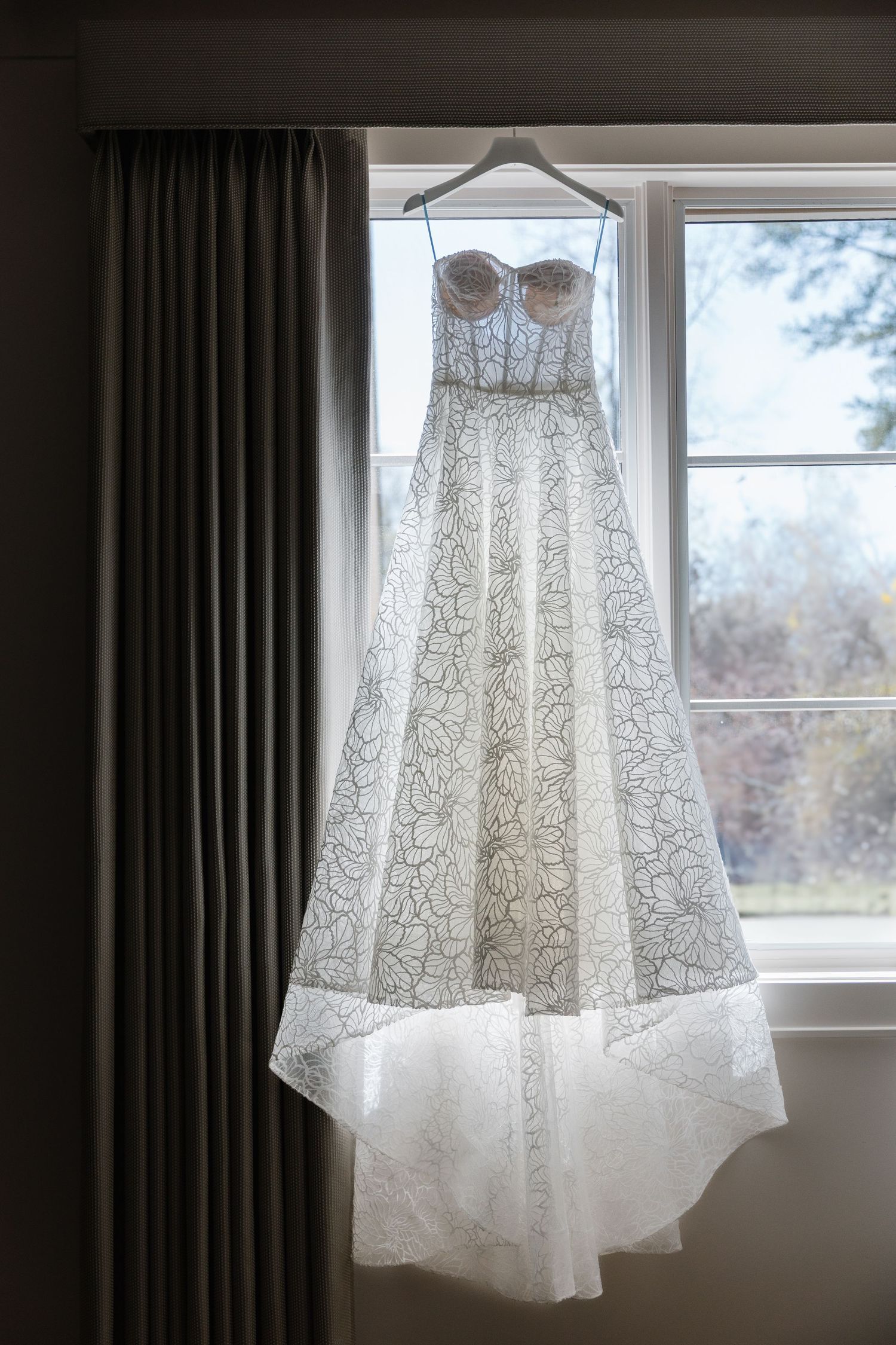 A white lace strapless wedding dress with floral patterns hangs in front of a sunny window on a padded hanger.