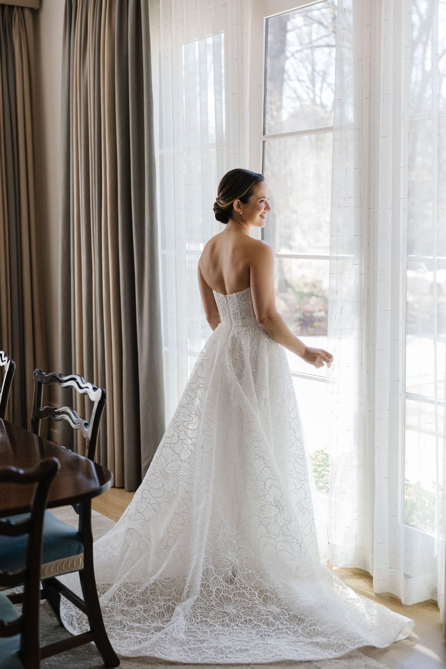 A bride in a stunning lace wedding gown with train gazes out a tall window beside elegant curtains and antique chair.