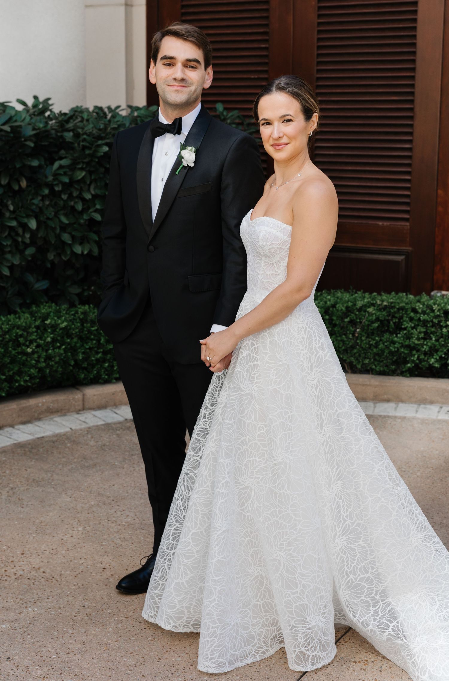 A bride in a white lace strapless gown and groom in classic black tuxedo pose together at their wedding.