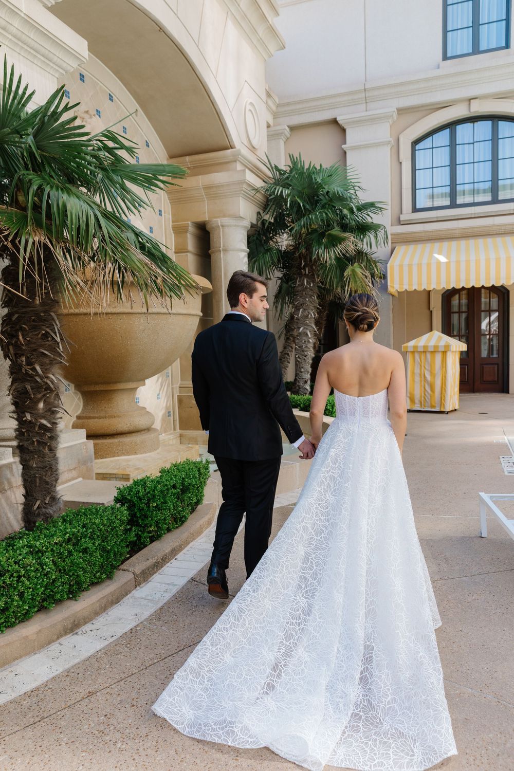 Elegant couple walking through luxurious archway with palm trees and striped awning during wedding celebration.