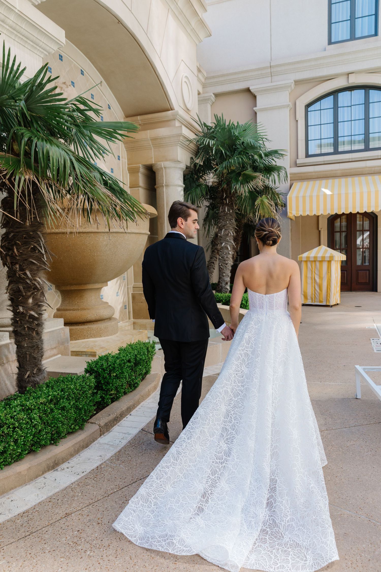Elegant couple walking through luxurious archway with palm trees and striped awning during wedding celebration.