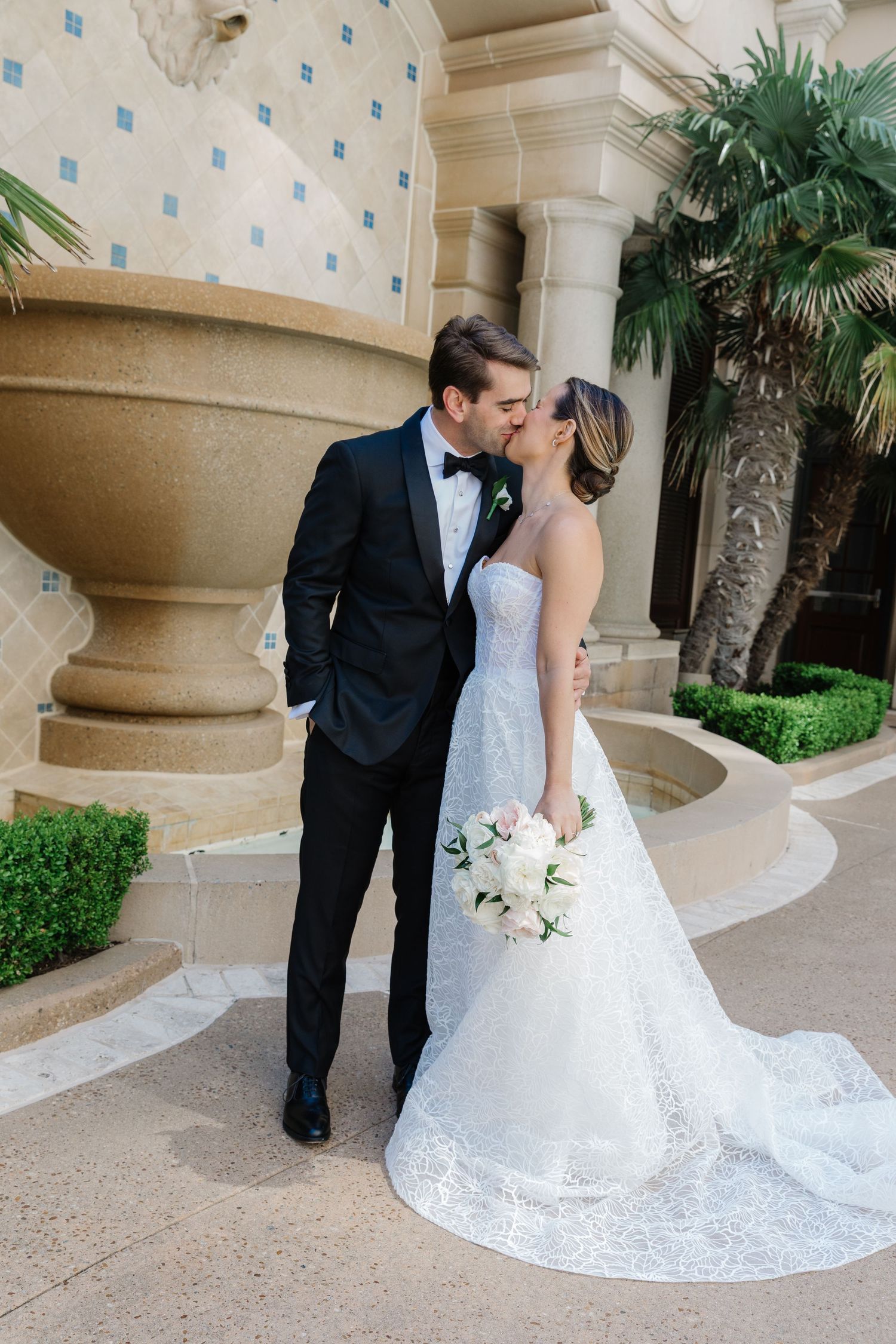 Newlywed couple sharing an intimate kiss in front of elegant stone columns and tropical palm plants.