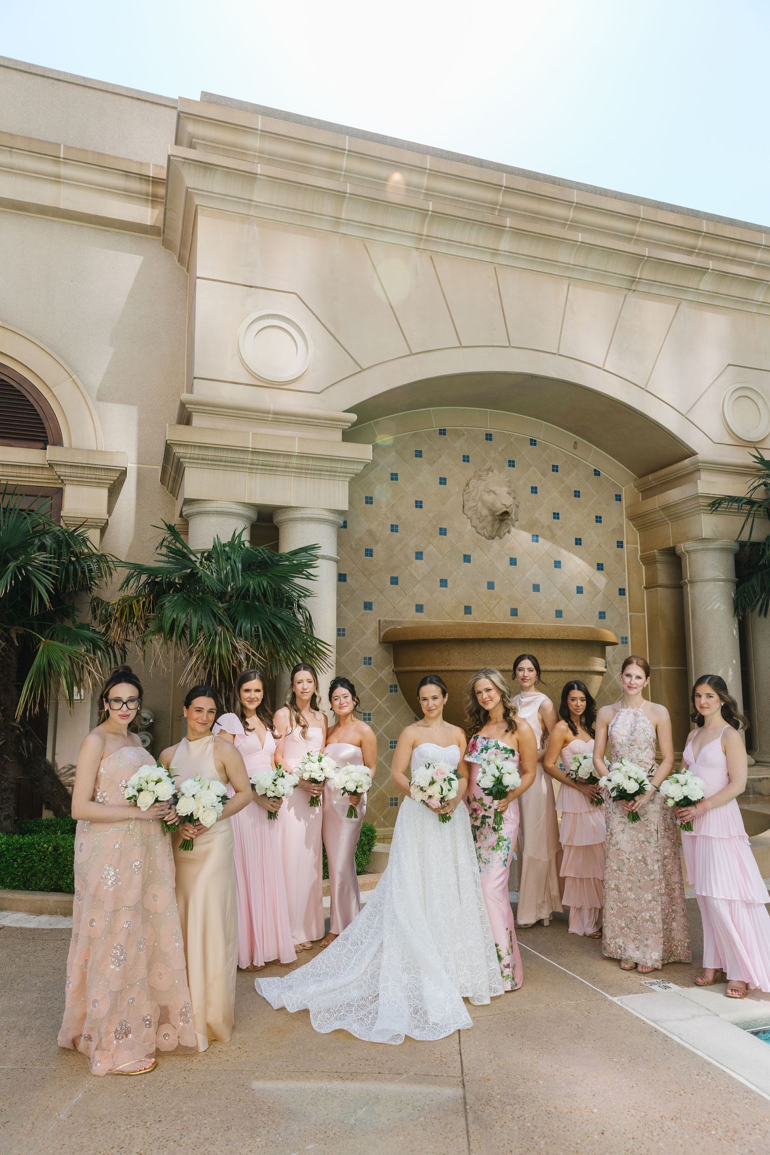 A wedding party in blush pink bridesmaid dresses and white bridal gown pose under an elegant arched entryway with columns.