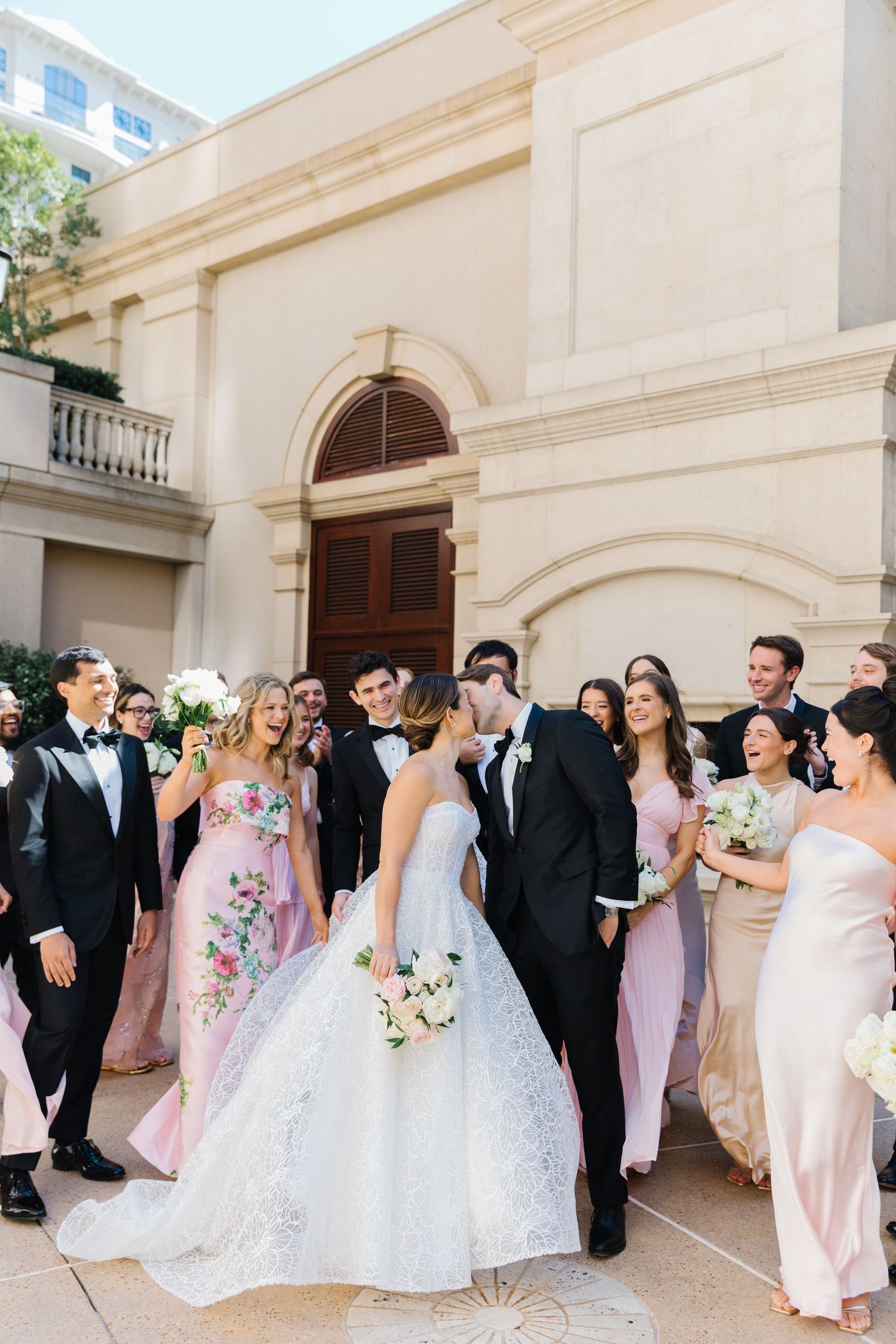 Wedding party gathers outside elegant cream-colored building with classical architecture for formal group photos.