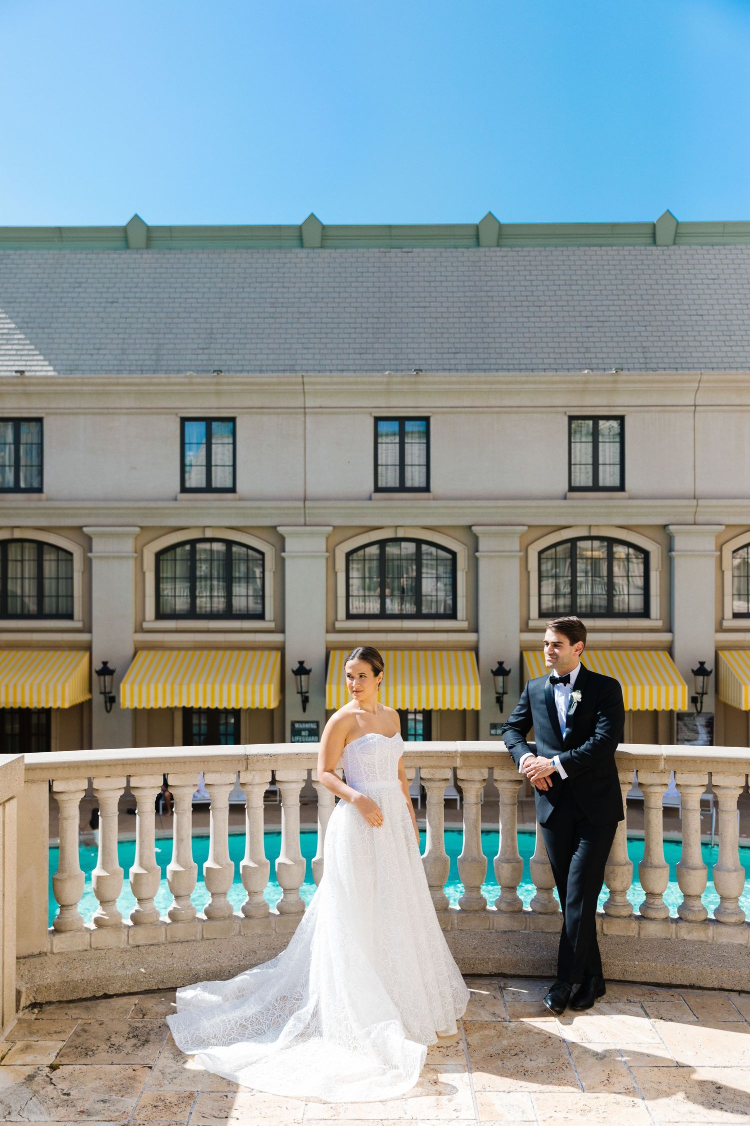 A couple in formal wedding attire stands on an elegant stone balcony with white columns against an upscale building facade.