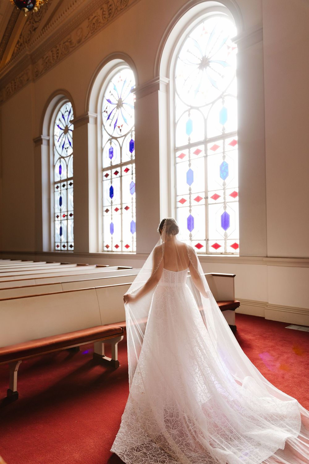 A flowing white wedding gown with trailing veil stands before ornate stained glass windows in a church with red carpet.