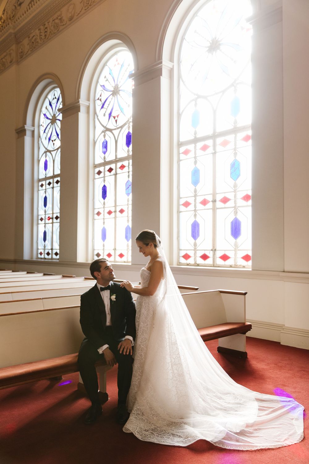A couple poses in a church before ornate stained glass windows, with a flowing white wedding dress on red carpet.
