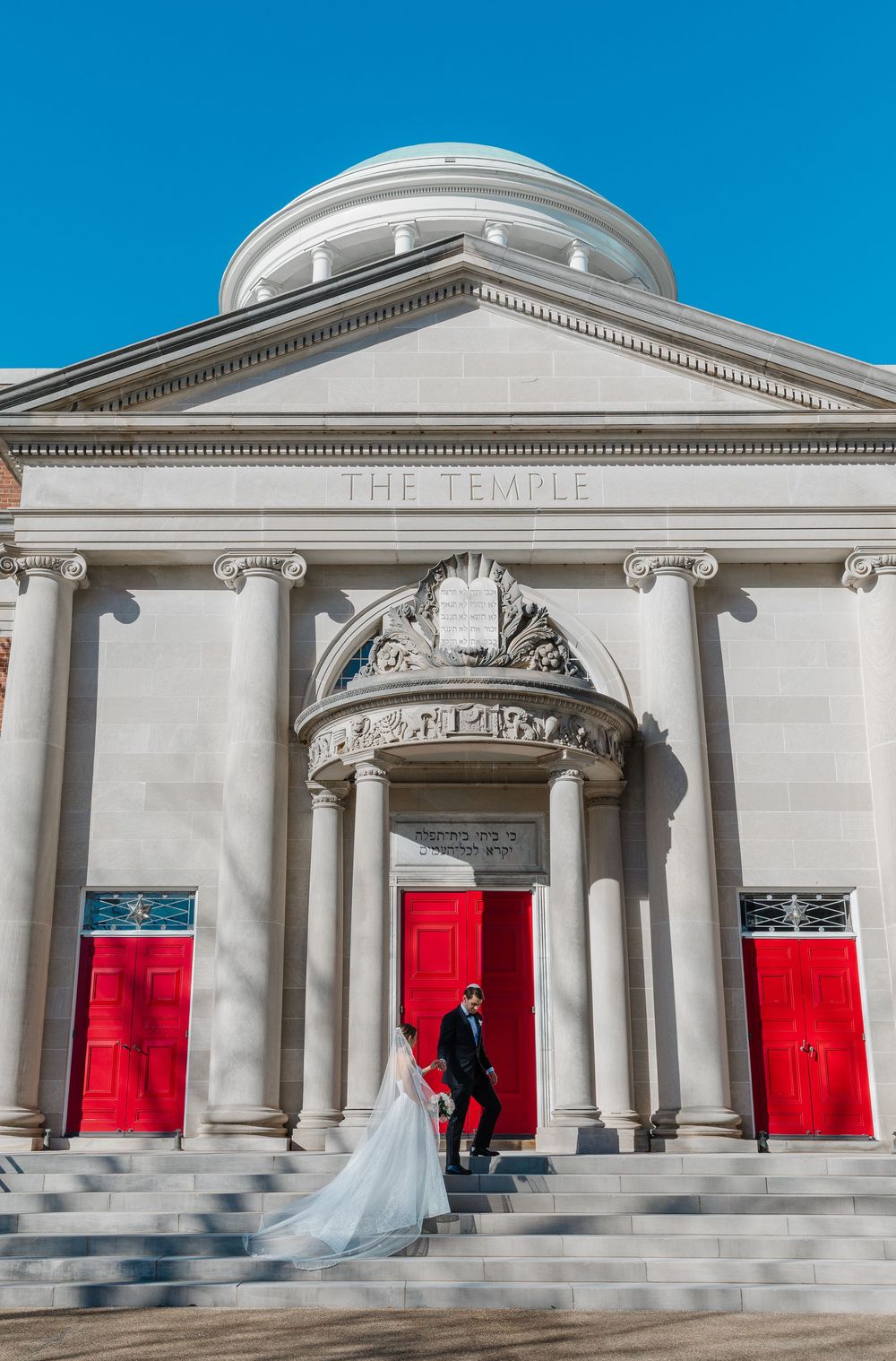 Elegant neoclassical church building with white columns and bright red doors against vibrant blue sky on wedding day.
