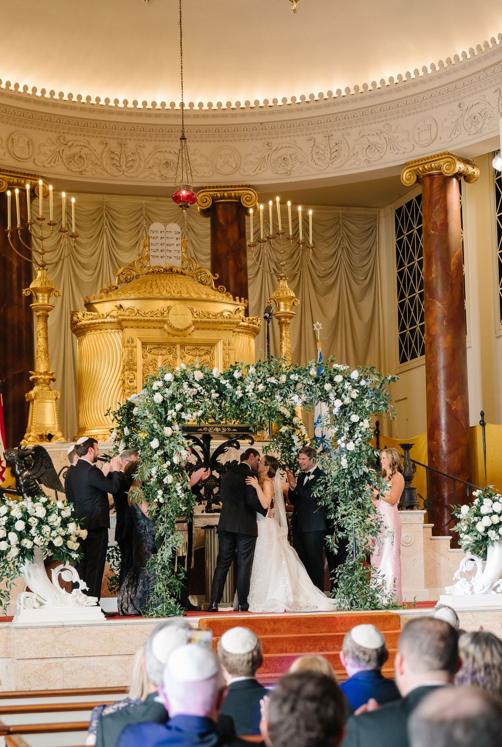 A wedding ceremony takes place under an ornate floral arch in a historic synagogue with elegant domed ceiling and candles.