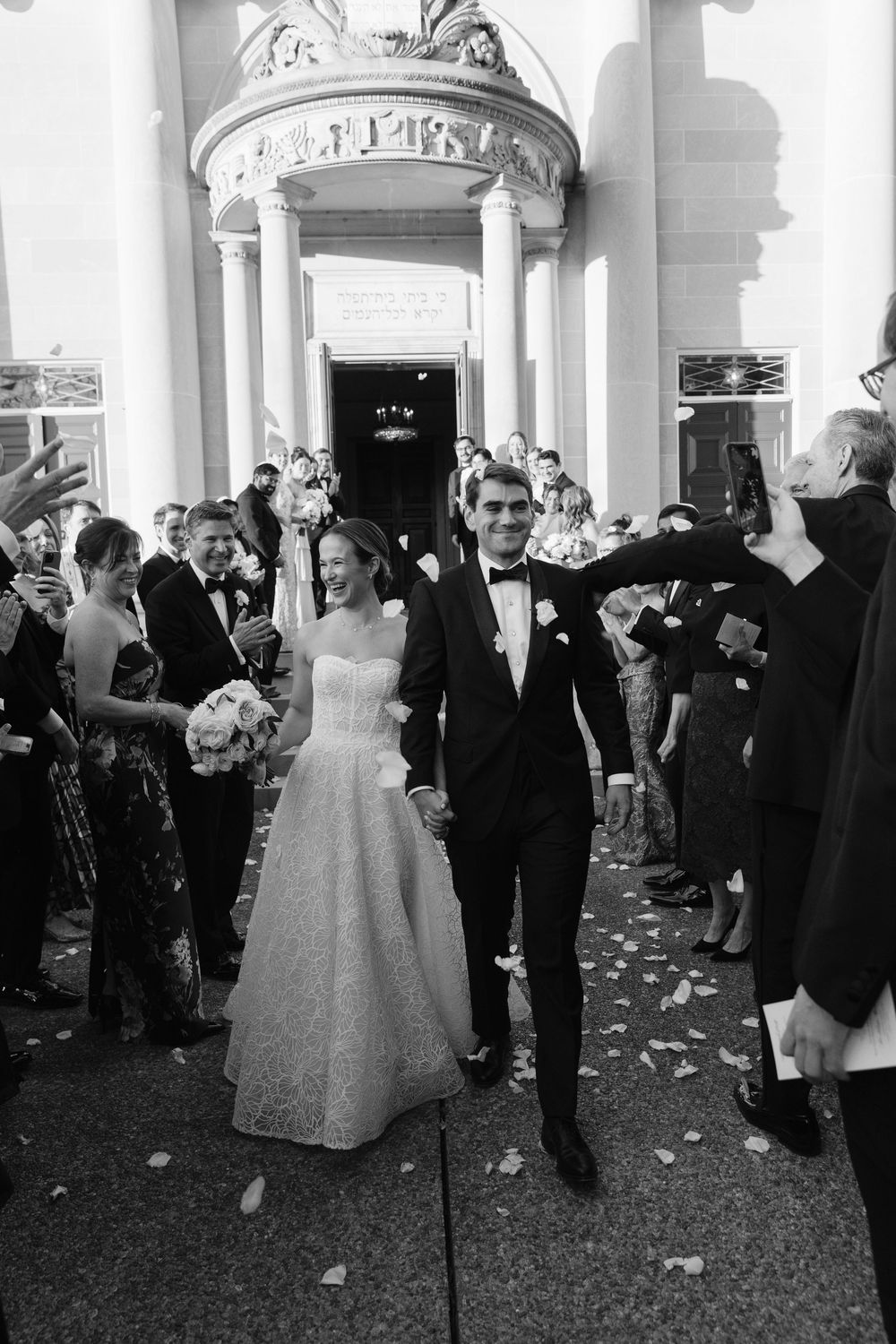 A couple exits their wedding ceremony as guests throw flower petals at an elegant columned entrance.