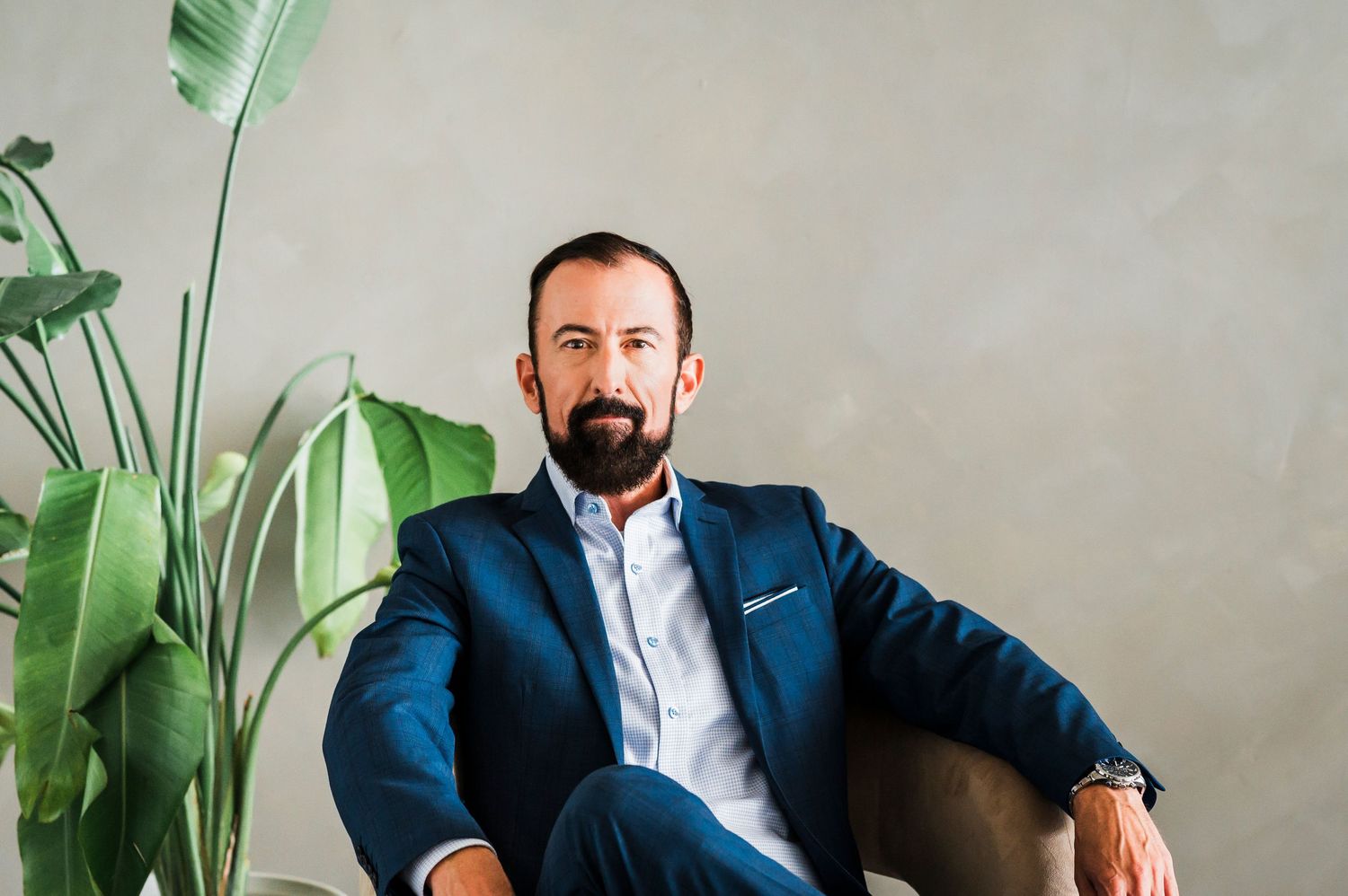 Professional dressed in a navy blazer sits casually in a chair next to a large green tropical plant against a gray wall.