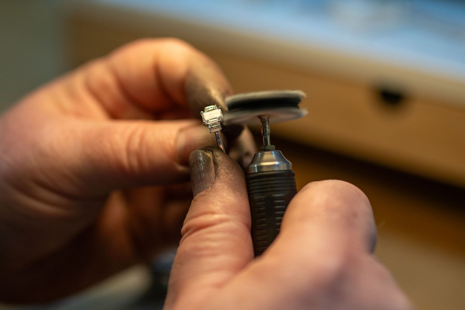 Hands examine a small diamond ring with jeweler's precision tools in a jewelry workshop setting.