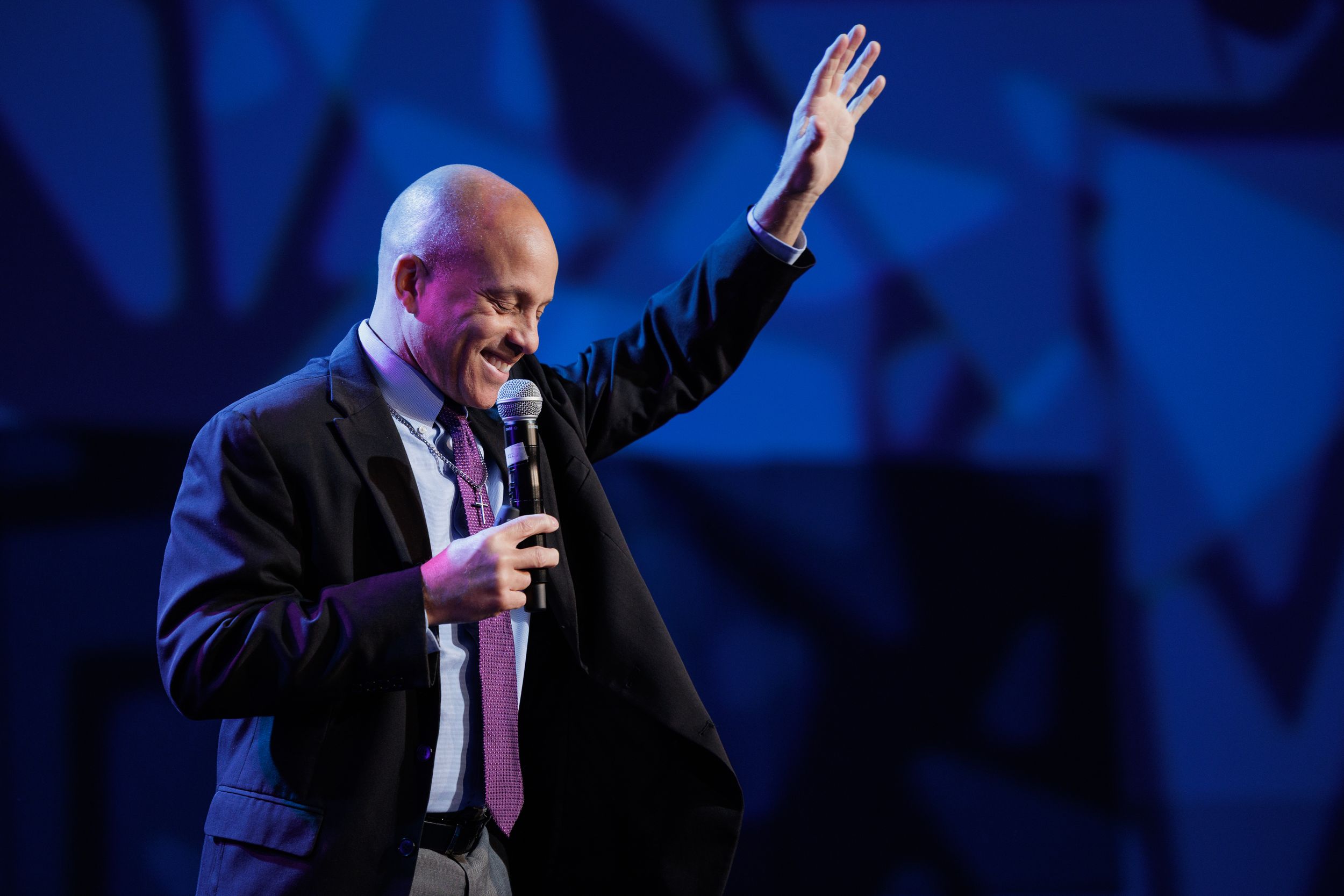 A speaker in a dark suit and pink tie gestures with one arm raised on stage with blue lighting behind them.