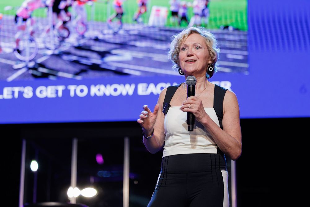 Person in a black and white outfit speaks on stage with colorful digital display in background at conference event.