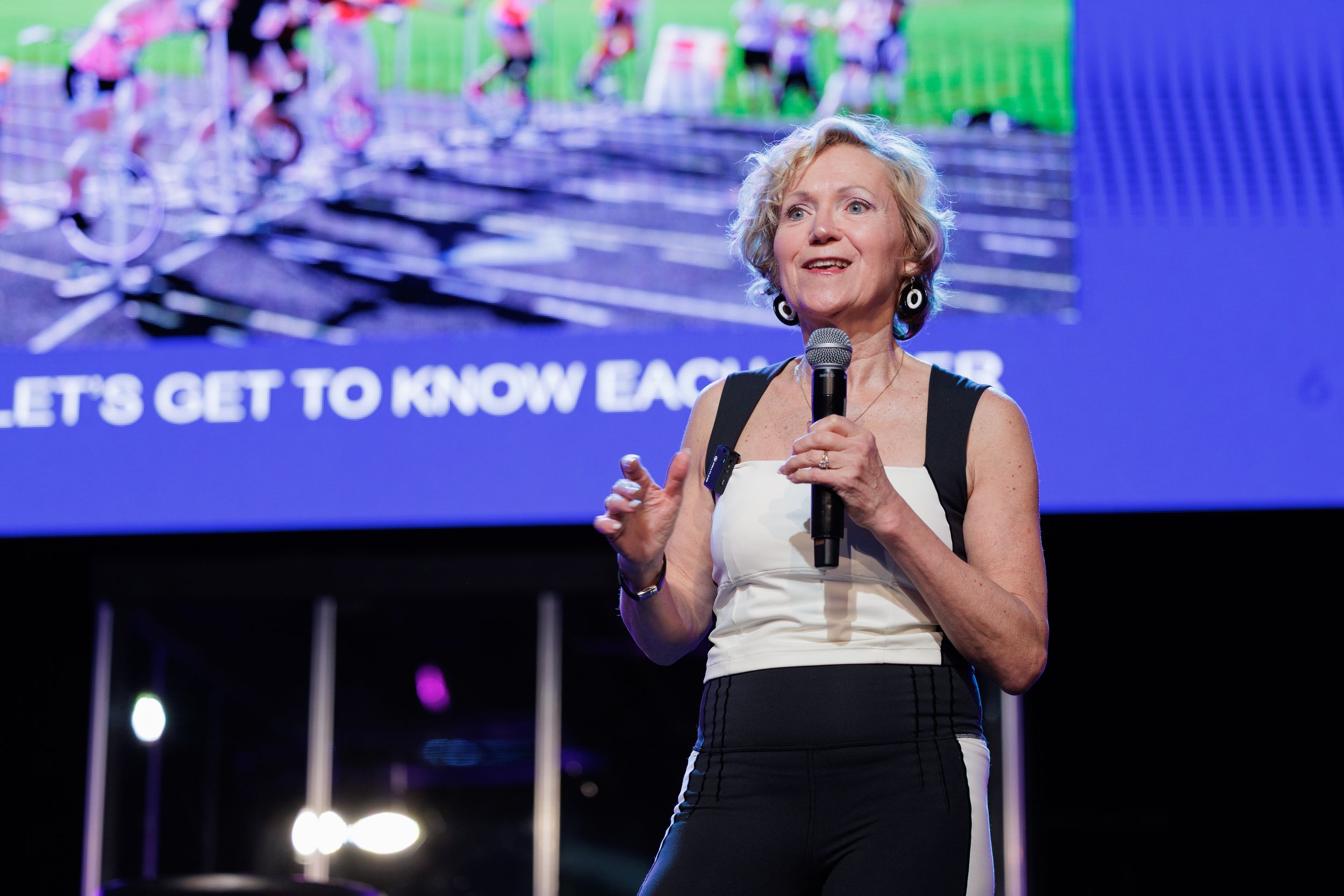 Person in a black and white outfit speaks on stage with colorful digital display in background at conference event.