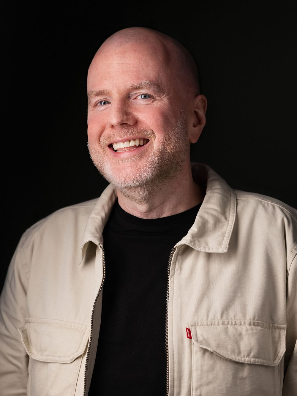 Professional headshot portrait of someone wearing a beige jacket over a black shirt against a dark background, smiling warmly.