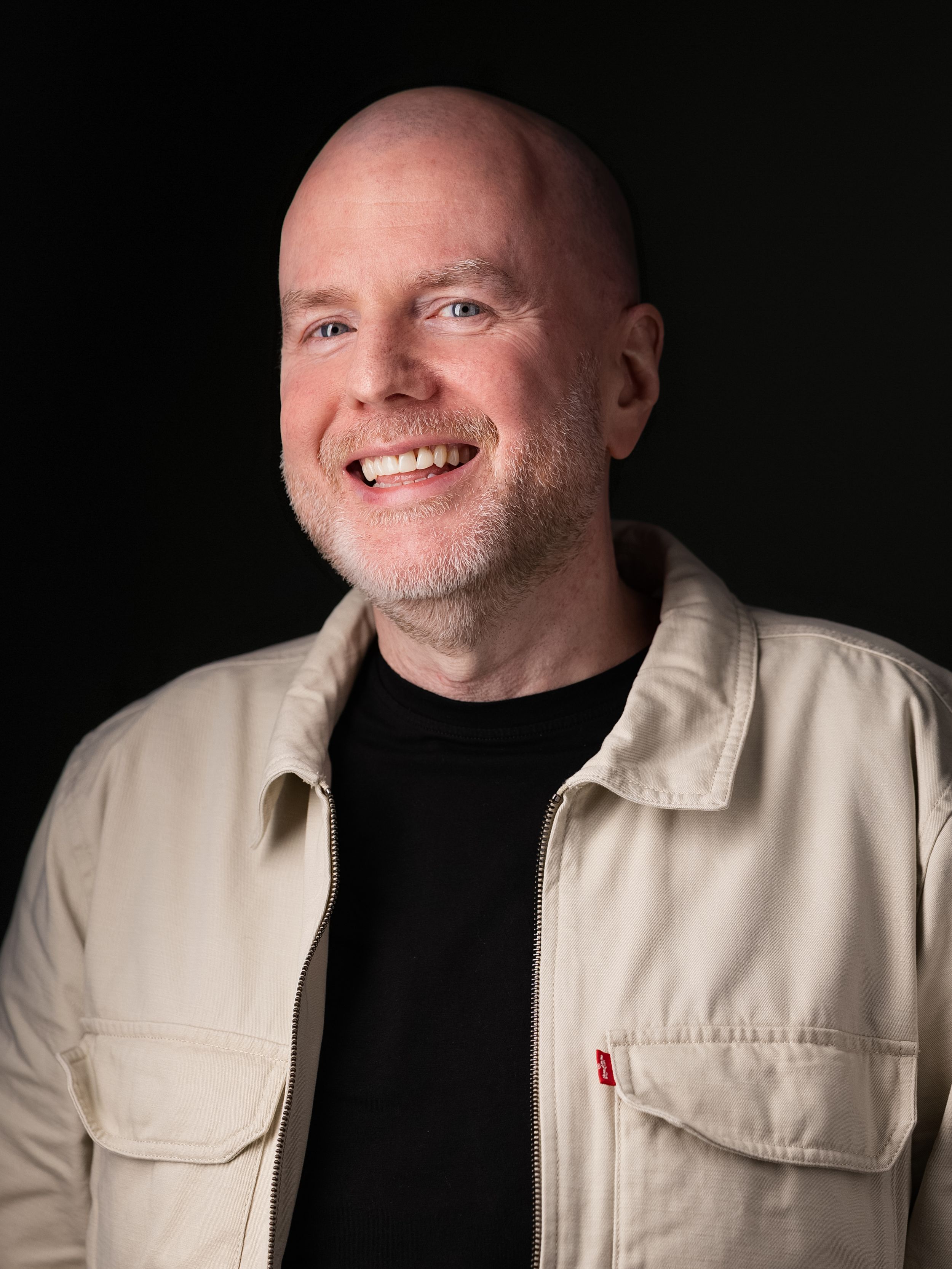 Professional headshot portrait of someone wearing a beige jacket over a black shirt against a dark background, smiling warmly.