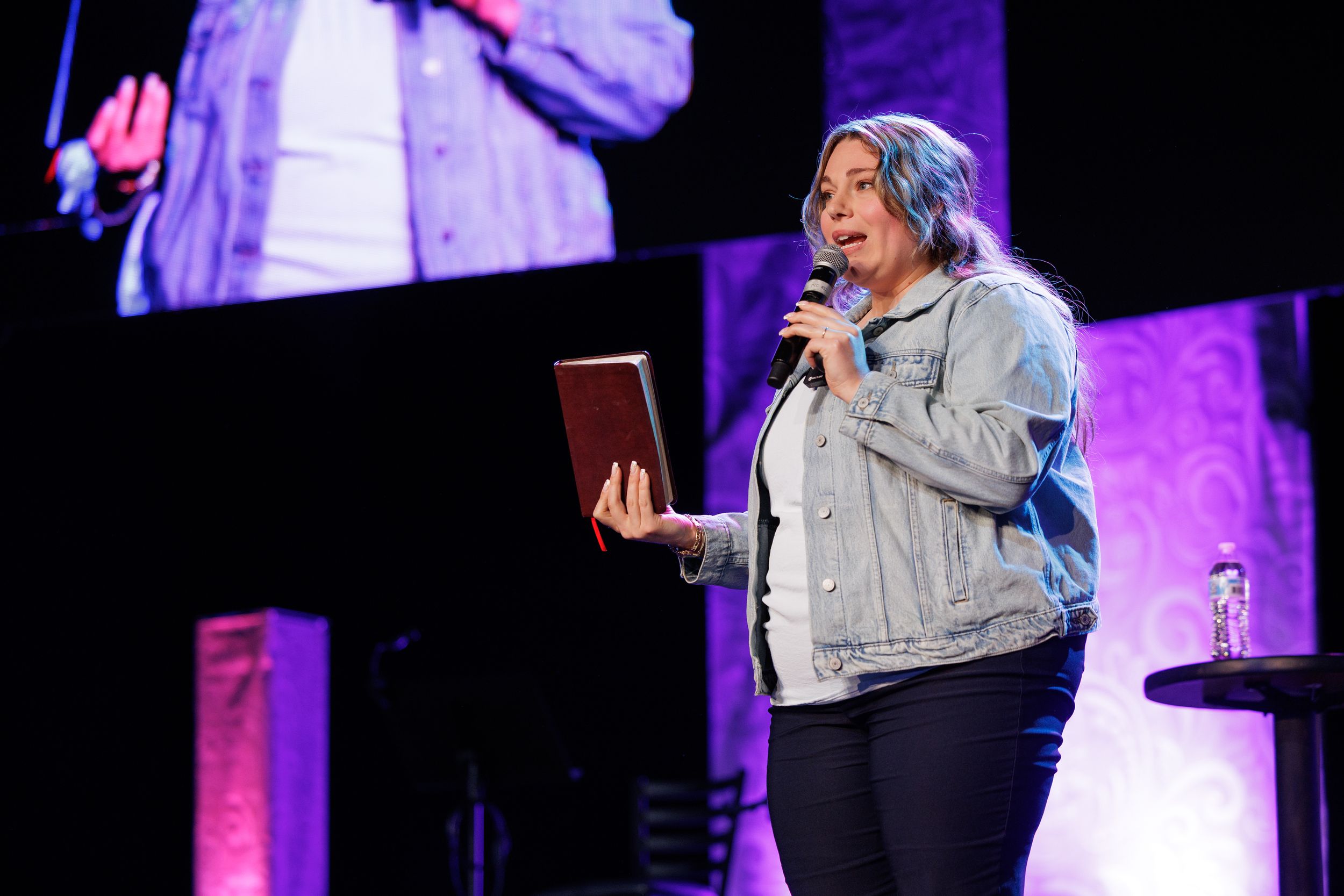 Speaker in light denim jacket holds a book on stage with large screens and purple lighting in the background.